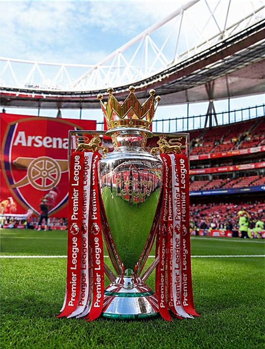 The image features a trophy, likely an Arsenal Premier League trophy, displayed prominently on a field at a stadium. The trophy is surrounded by red ribbons, which are often used as a symbol of victory or achievement. In the background, there's a large crowd gathered in the stands, indicating that this event has attracted fans and spectators from various locations. The image also includes text that reads "Trophies create history. Communities create momentum. Winning Arsenal is bringing football energy to the Solana ecosystem and the movement keeps getting stronger every day". This message emphasizes the importance of trophies as symbols of success and highlights how winning an event like this can contribute to the growth and development of a community or city, creating momentum for future achievements.