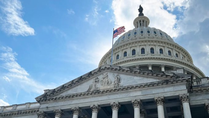 A low-angle view of the United States Capitol building under a partly cloudy sky, featuring its iconic white dome, columns, and an American flag flying above.