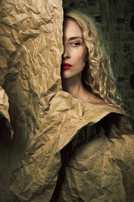 A blonde woman with red lipstick partially concealed behind crumpled brown paper, wearing a textured, draped garment in a moody setting.