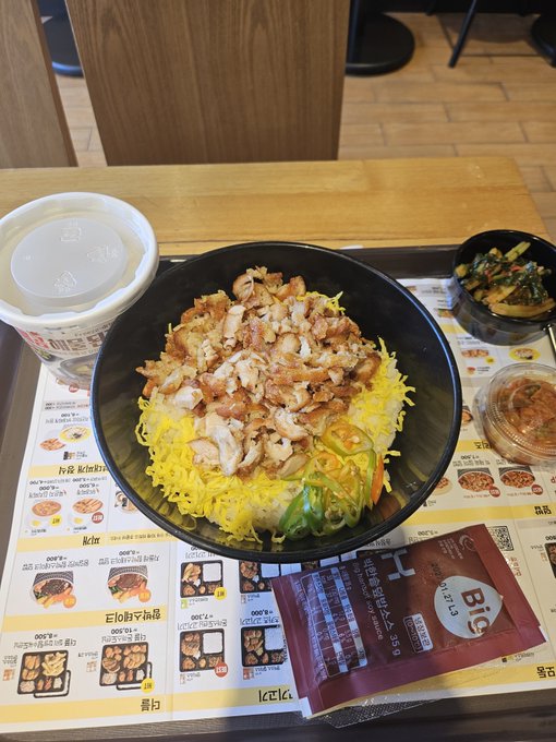 A black bowl filled with rice topped with grilled meat and shredded yellow egg sits on a tray menu, accompanied by a side dish of greens and a red packet labeled 'HOT SAUCE'.