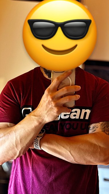 A person wearing a maroon t-shirt with a white smiley face emoji with sunglasses covering their head, taking a mirror selfie with a smartphone while wearing a white smartwatch.
