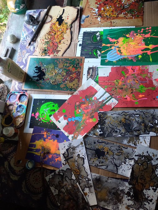 An overhead photograph displays a chaotic artist's workspace covered in various paint-splattered artworks, painting tools, and bottles of paint in an array of vibrant colors.