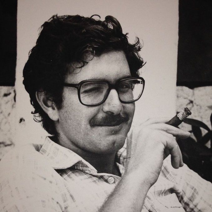 A black and white photograph of a smiling man with curly hair, a mustache, and large glasses, holding a cigar, appearing to be from an earlier era.