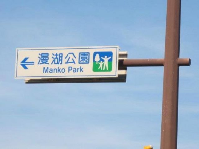 A directional street sign on a brown pole against a blue sky reading '漫湖公園 / Manko Park' with a left-pointing arrow and a park icon.