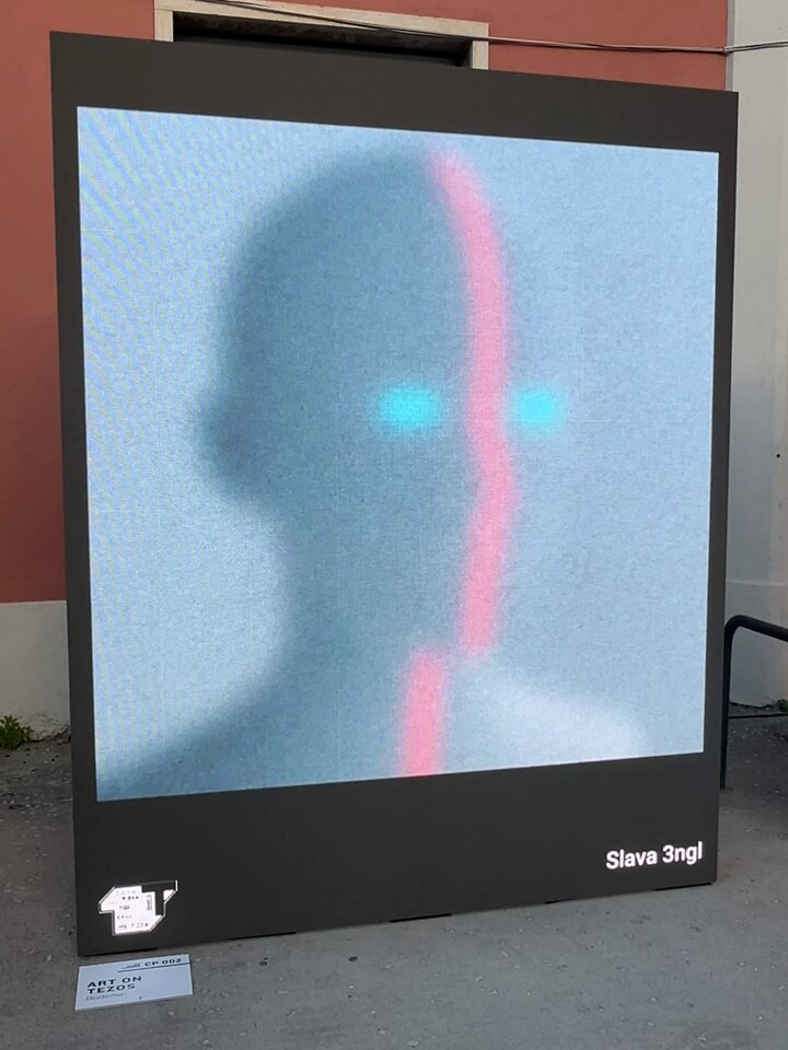 The image features a large screen or projector displaying an image of a person's face, with the name "Slava 3ngf" written below it. The projection appears to be a digital art piece that creates a shadow effect on the screen, giving the impression of a person's face being projected onto the wall. The background is a red brick building, which contrasts with the white wall where the projection takes place. The image also includes text that reads "Day 2 NFC Lisbon On view right at @NFCsummit for the Art on Tezos screening program: Cycles curated by @TeiaCommunity".