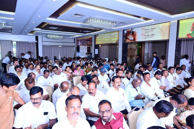 A large gathering of around 120 attendees, mostly men dressed in white shirts, sits attentively in a well-lit conference hall with chandeliers and banners on the walls, consistent with the Pi Network Karnataka conference held on September 28, 2026, to discuss education, GCV, and the Pi ecosystem.