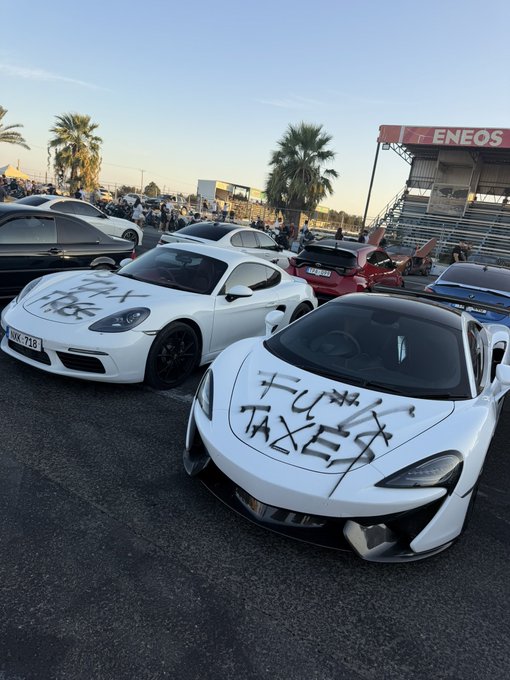 Two white sports cars—a Porsche 911 and a McLaren—are parked at what appears to be a car meet or racetrack (with an "ENEOS" banner visible), both tagged with graffiti reading "I FUKN TAXED" and "BKK-718" on the Porsche's plate. The image humorously illustrates the joke about non-domiciled residents quickly flaunting their tax-exempt wealth by purchasing exotic supercars.