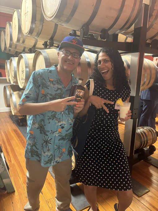 A man in a tropical Hawaiian shirt and a woman in a polka-dot dress smiling and holding drinks in a room filled with stacked wine barrels.