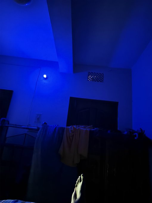 The image shows a person's foot with their toes pointing towards the camera, standing next to a black dresser or wardrobe that has clothes hanging out of it. The room appears dark, possibly due to the blue light from an overhead fixture. In the background, there is a bed and a TV mounted on the wall. The post features a caption about a person's foot being in the foreground while others sleep, suggesting that someone might be watching or recording something happening at night.