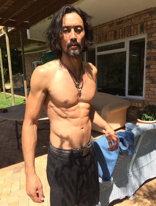 A shirtless, lean man with long dark hair and a beard stands on a patio, wearing dark pants and a pendant necklace.