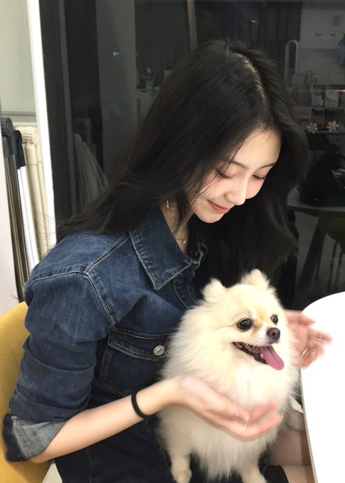A young woman with long black hair, wearing a denim jacket, smiles down at a fluffy white Pomeranian dog she is holding.