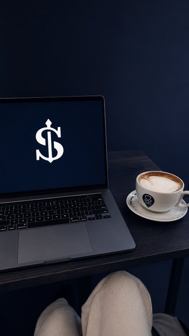A laptop displaying a white dollar sign logo on a dark screen sits on a wooden table next to a cappuccino in a white cup with a matching logo, with a person's lap visible in the foreground.