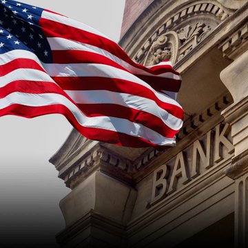 The image features a large American flag flying above an old, historic building that has the word "Bank" written on its facade. The flag is waving in the wind, creating a sense of patriotism and pride for the country. The bank building itself appears to be quite old and grand, with intricate architectural details. The post accompanying this image discusses the importance of community banks and credit unions utilizing blockchain technology to stay competitive and relevant in today's rapidly changing financial landscape.