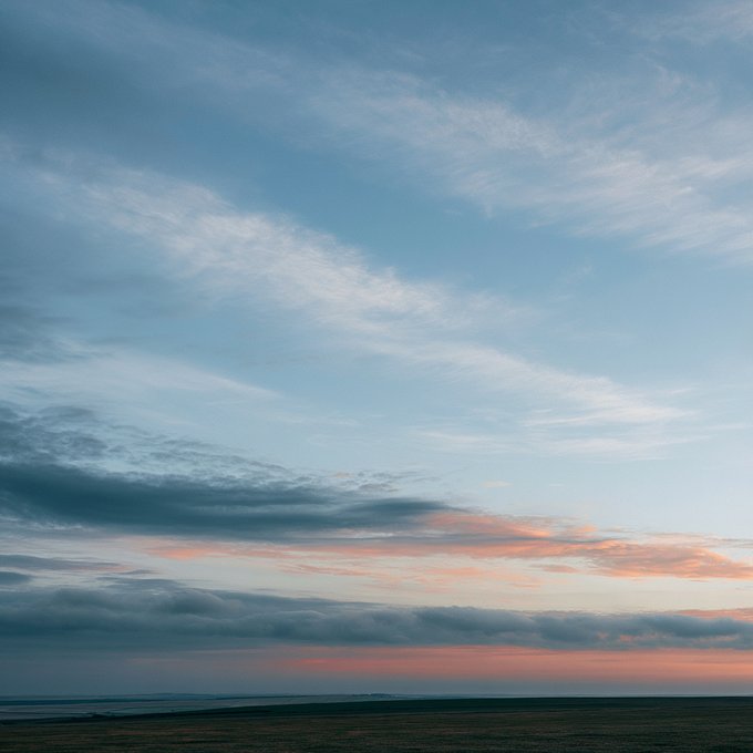 A serene landscape photograph showing a sunset or sunrise over a dark horizon with wispy clouds tinted in pink and orange against a blue sky.