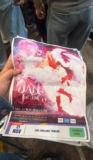 A hand holds a fan-made promotional flyer for 'Jan Jingjing Fancon' dated Saturday, 21 November 2026, at Thunderdome, featuring anime-style artwork of a long-haired figure and listing various event prices and inclusions.