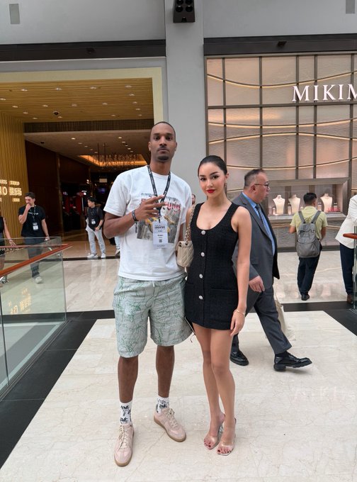 A tall man wearing a "$GOODY" lanyard stands next to a young woman in a black button-down dress inside a luxury mall, with "MIKIMOTO" visible in the background. The image humorously supports the caption by implying the woman's impressive height is the reason the poster hasn't sold their $ANSEM bags, as her towering presence suggests significant gains.