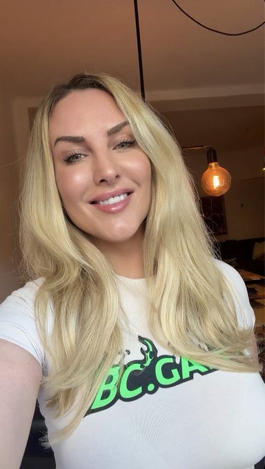 A woman with long blonde hair smiles at the camera while wearing a white t-shirt with green 'BC.GA' lettering, standing in a room with an exposed bulb pendant light.