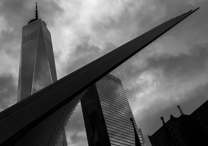 A dramatic black-and-white photo captures a low-angle view of the One World Trade Center tower alongside a modern spired structure, set against a cloudy sky.