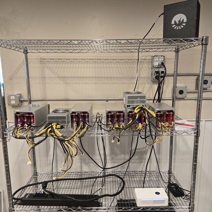 A metal shelving unit holds multiple cryptocurrency mining rigs with red indicator lights, yellow power cables, and control boxes mounted on the wall.