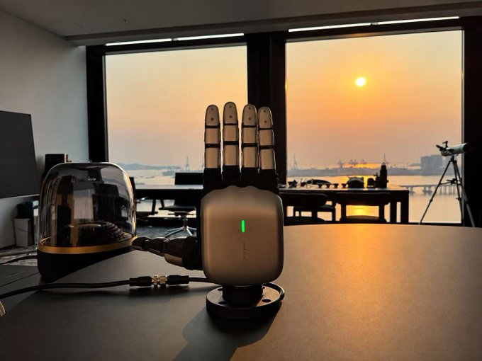 A small metallic desktop device labeled "LEADIN" with four antenna-like projections sits on a desk in front of a window showing a sunset over a harbor, with a glass cloche and telescope visible nearby.