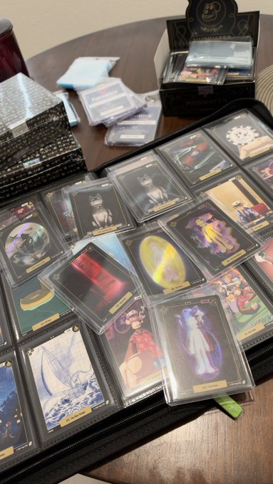 A collection of Pokémon trading cards in protective sleeves is spread across a table and open card binder, with more cards and storage supplies visible nearby.