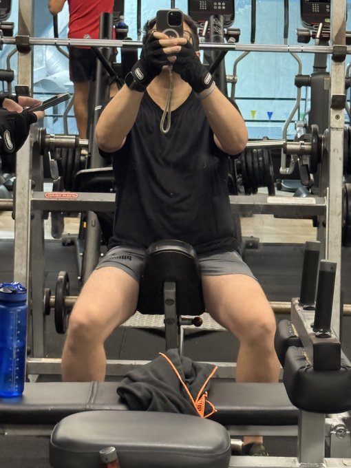 A person wearing black workout gloves and shorts takes a mirror selfie at a gym using a smartphone, sitting on a weight machine with a blue water bottle nearby.