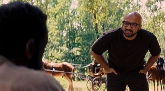 A bald man with glasses and a black t-shirt stands with his hands on his hips, looking at another person in the foreground, with horses visible in a wooded background.