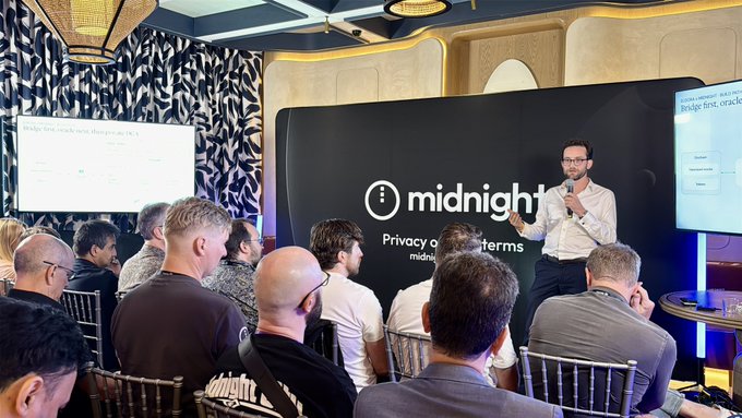 A man in a white shirt and glasses speaks to a seated audience while holding a microphone, with a 'midnight' banner behind him and a presentation slide titled 'Bridge first, evolve later' on a screen to the left.