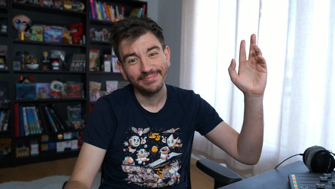 A man in a black graphic T-shirt smiles and makes an 'OK' hand gesture while sitting in a room with shelves of video games and figurines behind him.