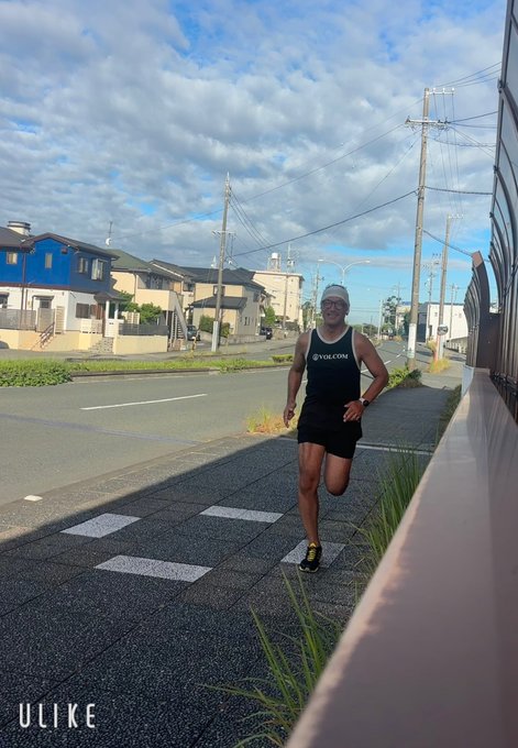 A runner wearing a black "u vlcome" tank top, shorts, and white cap jogging along a sidewalk in a residential area under a partly cloudy sky.