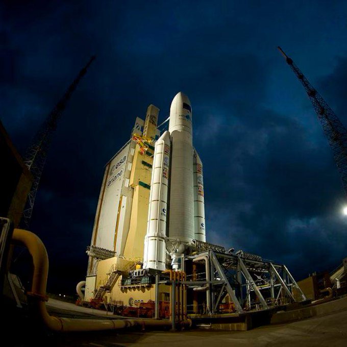 In this image, a large rocket ship is being prepared for launch against a dark sky with clouds. The rocket is situated near a building that has two towers on its roof, which could be related to the launch facility or another structure nearby. The scene captures the anticipation and excitement of launching a space shuttle into space.