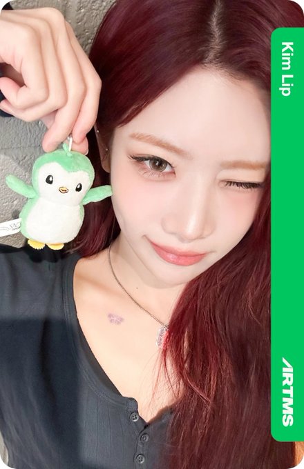 A selfie of a woman with red hair winking and holding a green penguin plush keychain, with 'Kim Lip' and '/RTMS' text visible on a green sidebar.