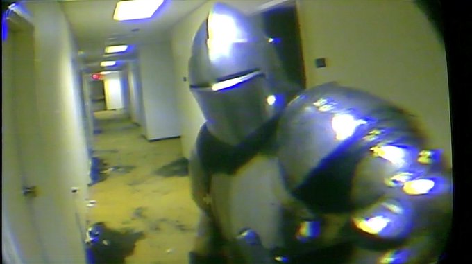 A distorted, low-quality image shows a person in silver armor standing in a hallway, filmed through a peephole or fisheye lens, with light reflecting off their helmet.