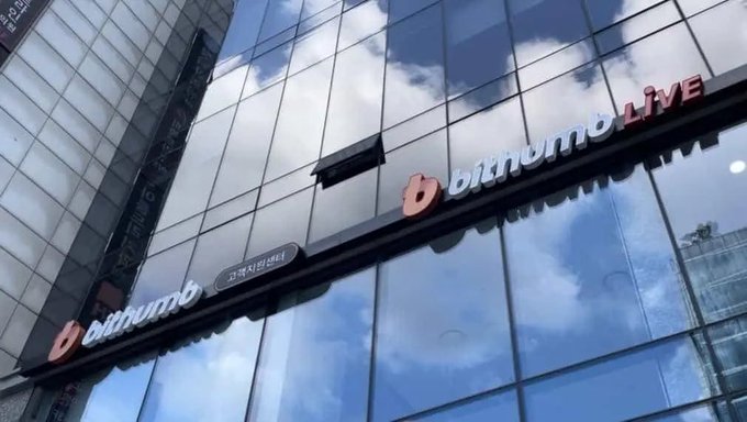 The exterior of a glass-fronted building displays a sign reading "bithumb LIVE" with cloud reflections visible in the windows.