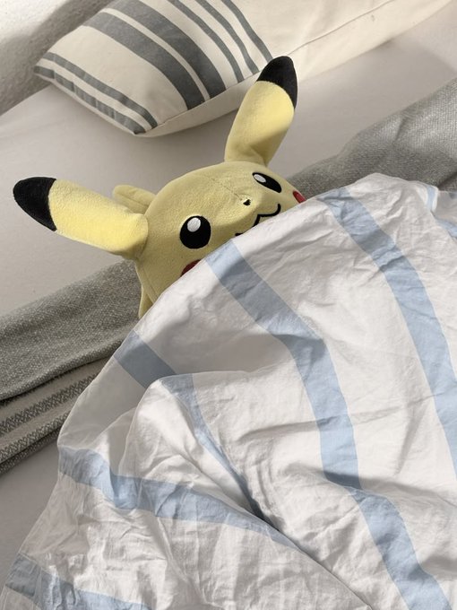 A Pikachu plush toy peeking out from under a blue and white striped blanket on a bed with pillows.