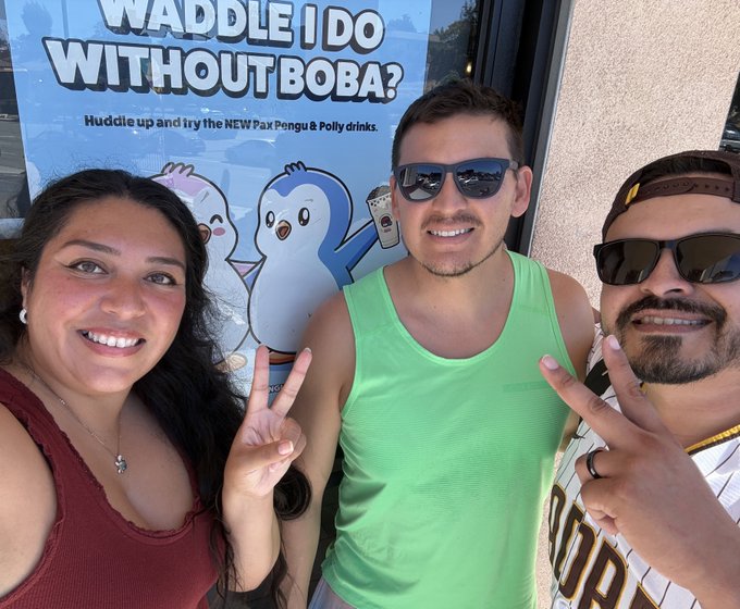 Three smiling people pose for a photo outside a boba shop, with two making peace signs in front of a window decal reading 'WADDLE I DO WITHOUT BOBA? Huddle up and try the NEW Pax Pengu & Polly drinks.'
