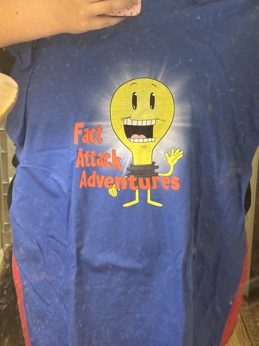 A blue t-shirt features a smiling yellow lightbulb character alongside the text "Fact Attack Adventures," excitedly being held up as the new merchandise arrives.