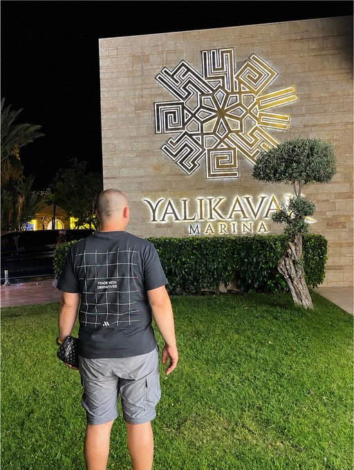 A man wearing a gray "Track Mile Clothes" t-shirt stands in front of the illuminated Yalıkavak Marina sign, which features a large geometric emblem, during the evening. The image reflects the post's message of spending a pleasant evening at Yalıkavak Marina while showcasing the gift t-shirt from Midas.
