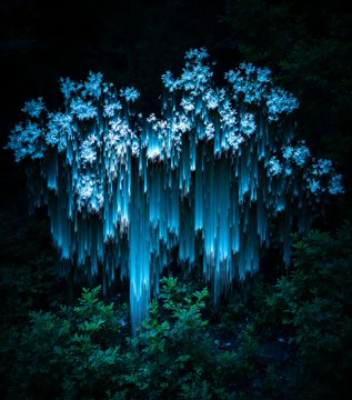An ethereal, glowing tree of light blue energy stands in a dark forest, with luminescent branches and cascading tendrils that resemble hanging vines or falling water.