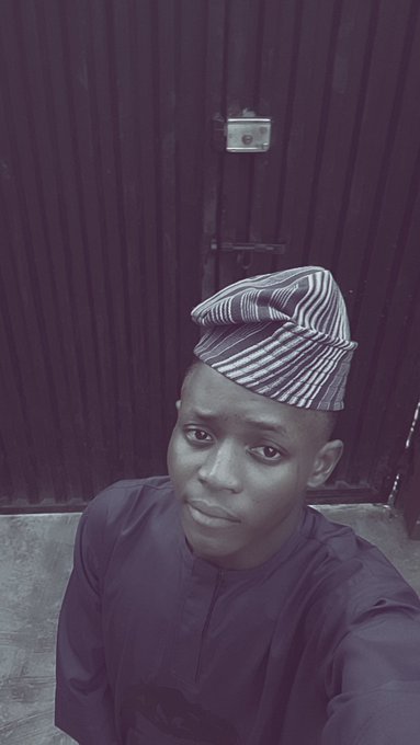 A selfie of a young man wearing a traditional patterned cap and a dark traditional outfit, standing in front of a dark wooden door.