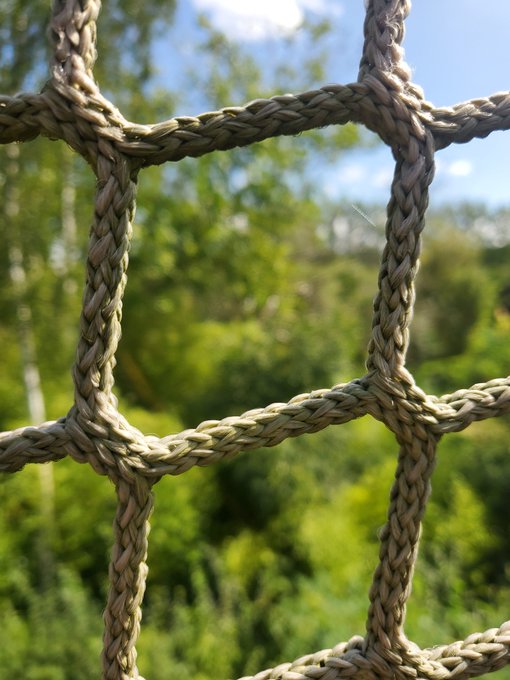 A close-up view of a thick, braided rope net with green foliage and blue sky visible in the blurred background.