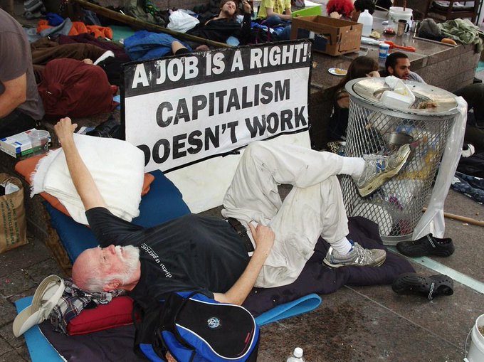 A man with a white beard and black 'people for bernie' t-shirt lying on the ground next to a sign reading 'A JOB IS A RIGHT! CAPITALISM DOESN'T WORK', surrounded by belongings and other people in what appears to be a protest encampment.