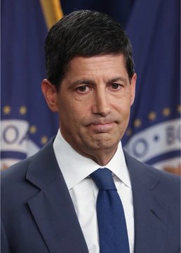 A serious-looking man in a dark blue suit and tie stands in front of what appears to be an official government backdrop with gold fringe and stars, suggesting a press or official setting. The image relates to the post by showing the Federal Reserve official who is anticipated to potentially raise interest rates, with the caption warning viewers to prepare for market dips.