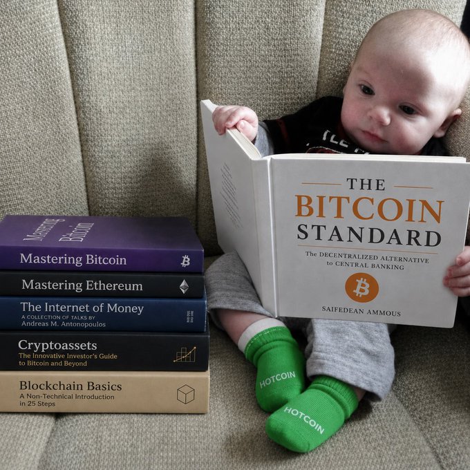 A baby wearing green 'HOTCOIN' socks sits reading 'The Bitcoin Standard' by Saifedean Ammous, with a stack of crypto books beside them including 'Mastering Bitcoin,' 'Mastering Ethereum,' 'The Internet of Money,' 'Cryptoassets,' and 'Blockchain Basics.'