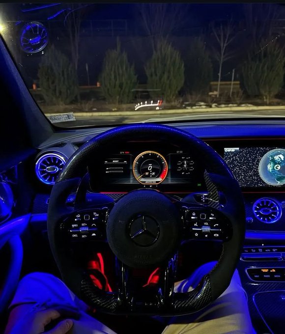 Interior view of a Mercedes-Benz cockpit at night, featuring a flat-bottomed steering wheel, digital instrument cluster, and ambient blue and red lighting.