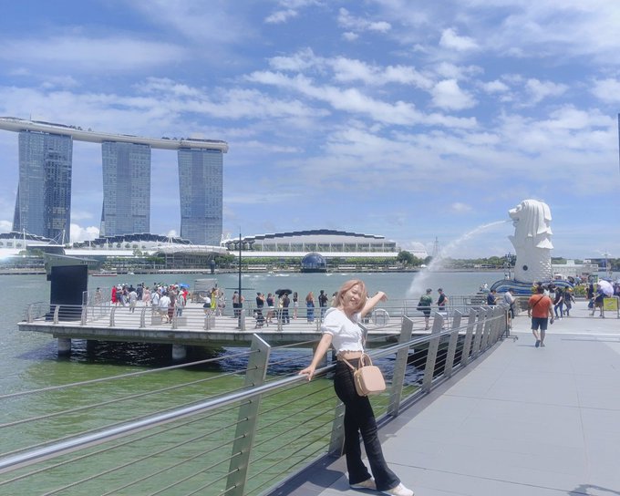 A woman in a white shirt and black pants poses for a photo on a waterfront promenade in Singapore, with the Merlion statue, Marina Bay Sands hotel, and ArtScience Museum visible in the background under a partly cloudy sky.
