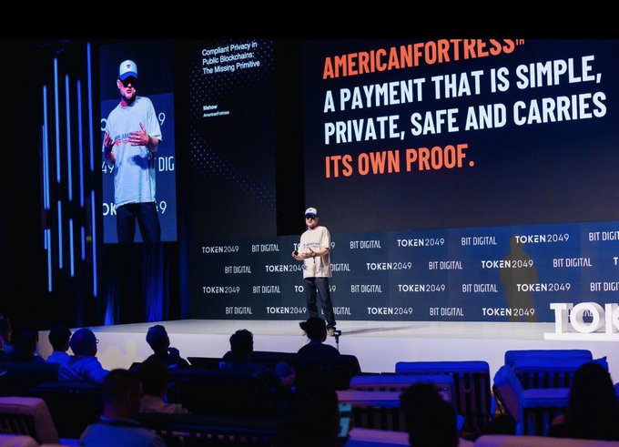 A speaker presents at the TOKEN2049 BIT DIGITAL conference, with on-screen text highlighting "Compliant Privacy in Public Blockchains: The Missing Primitive" and an "AMERICANFORTRESS" tagline describing it as "A PAYMENT THAT IS SIMPLE, PRIVATE, SAFE AND CARRIES ITS OWN PROOF." The presentation aligns with the post's discussion of how AmericanFortress addresses blockchain payment transparency by offering a privacy solution that maintains regulatory compliance.