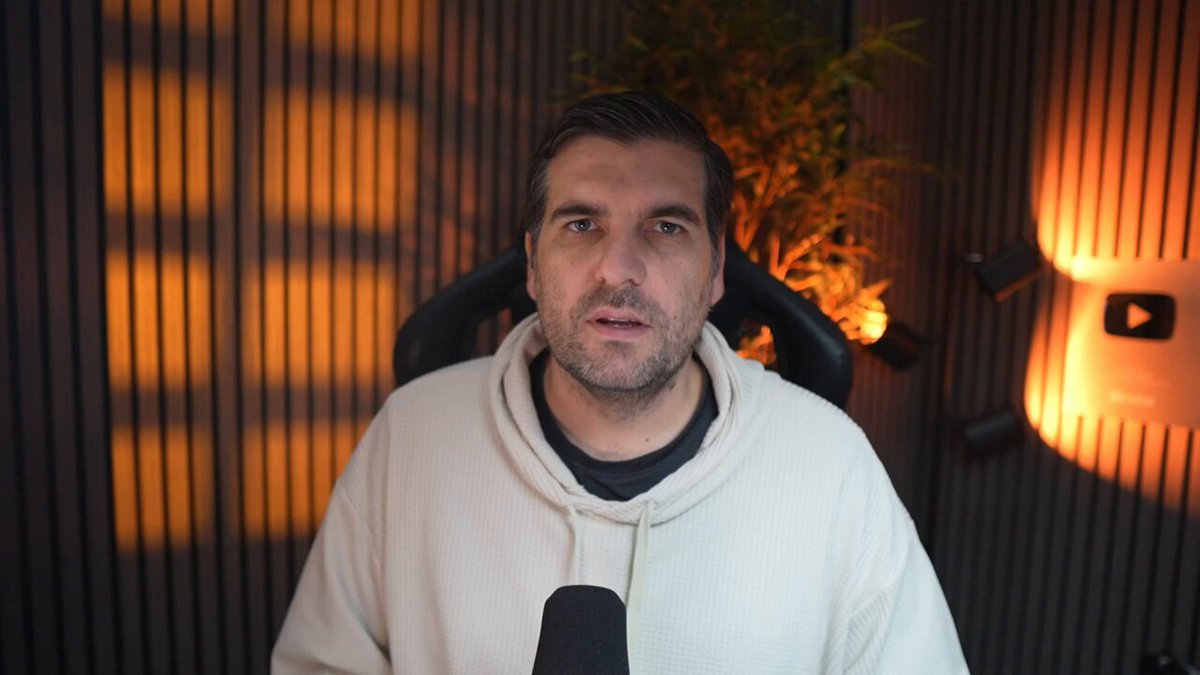 The image shows a man wearing a white hoodie sitting behind a microphone with his hands on his lap. The background features a dark wall with orange lights shining through the slats, creating an interesting visual effect. The image also contains text that reads "Coinbase data reveals a 7x XRP anomaly... Unexplained Coinbase data reveals a startling seven to one buy liquidity imbalance for XRP". This suggests that there is some unusual or unexpected information related to cryptocurrency trading on Coinbase.