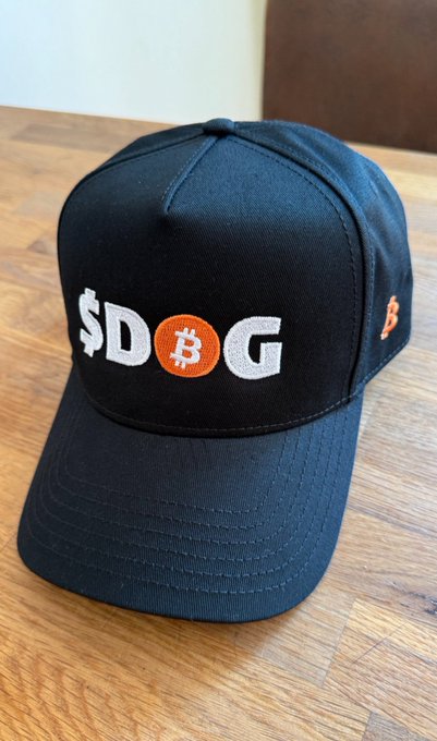 A black snapback cap features the embroidered text "$DOG," with the "O" replaced by an orange Bitcoin "₿" symbol, sitting on a wooden surface. The hat's design ties into the "$DOG ARMY IS DIFFERENT" post by blending meme culture with crypto branding, appealing to the playful, community-driven spirit of the Dog Army.
