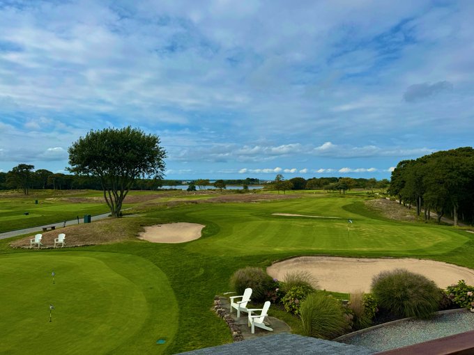 An aerial view of a golf course with sand bunkers, putting greens, flagsticks, and a few white lounge chairs, with trees and water visible in the distance under a partly cloudy sky.